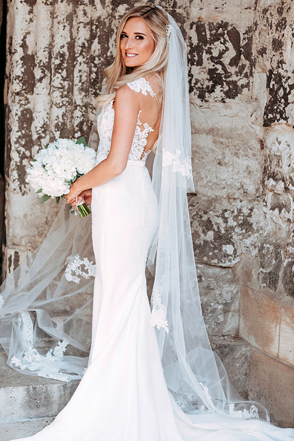 A bride in a fitted gown and long veil holding white roses